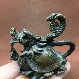 Puede incluir: Figura de bronce que representa una criatura mítica con cabeza de dragón y cuerpo de tortuga. Una serpiente se arquea por encima, sosteniendo una moneda. La figura está detallada con escamas.