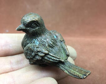 May include: A small, bronze-colored bird figurine with detailed feather textures. The bird has a slightly weathered appearance, with hints of blue-green patina. The figurine is resting on a hand, showcasing its size and intricate design.