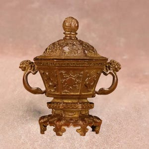Puede incluir: Un quemador de incienso decorativo de color bronce con tapa, tallas ornamentadas y dos asas con cabeza de león. El quemador se asienta sobre una base decorativa con un diseño detallado. La tapa está rematada con un adorno decorativo.