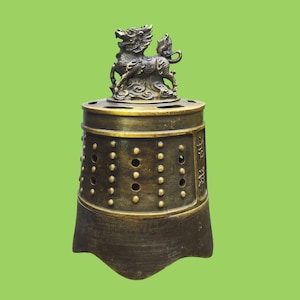 May include: A dark bronze-toned metal incense burner shaped like a traditional bell. The top features a sculpted mythical creature figure. The cylindrical body is decorated with rows of raised circular studs and vertical panels with engraved text.
