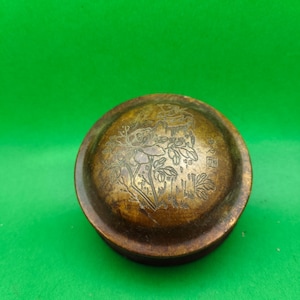 May include: A circular, antique bronze box featuring an intricate floral and architectural pattern on its lid. The box has a slightly aged appearance, indicating its history. The design is etched into the metal, providing a textured surface.