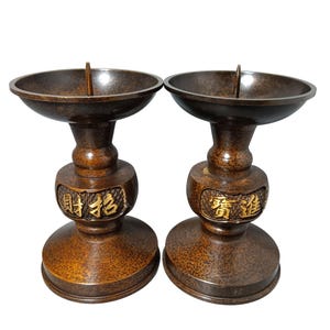 May include: Two antique bronze candle holders. Each has a round top, a pedestal base, and gold Chinese characters. The candle holders are brown with a textured surface and a small metal spike in the centre.