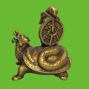 Pode incluir: Uma estatueta de metal cor de latão representando uma criatura mítica com cabeça de dragão e corpo de tartaruga, com uma cobra enrolada nas costas segurando uma moeda circular com caracteres em relevo e uma pequena criatura no topo.