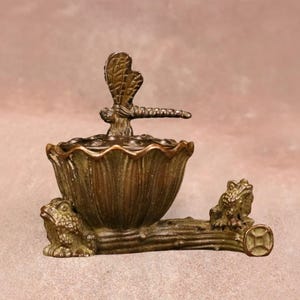 Puede incluir: Quemador de incienso decorativo de bronce con una libélula. El quemador tiene forma de flor y está sostenido por un tronco con figuras de ranas. Un artículo único para la decoración del hogar.