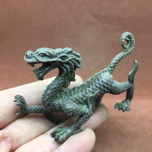 Può includere: Statuetta di drago in bronzo dettagliata con corpo strutturato e squamoso e bocca aperta. Il drago ha una coda arricciata ed è posato con le zampe anteriori estese. La statuetta è di colore verde scuro.