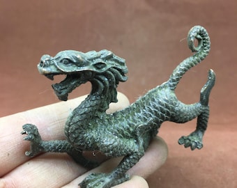 May include: A detailed bronze dragon figurine with a textured, scaled body and open mouth. The dragon has a curled tail and is posed with its front legs extended. The figurine is a dark green color.