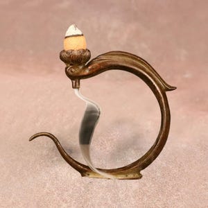 May include: A bronze-colored incense holder with a decorative, curved design. The holder features a floral base supporting a cone-shaped incense stick with a white tip. Smoke gently rises from the incense, creating a visual effect.