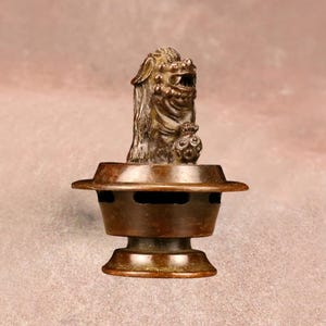 Puede incluir: Un quemador de incienso de bronce con una figura de guardián con forma de león. El quemador tiene un diseño escalonado con orificios de ventilación. El color general es un bronce cálido y envejecido. El diseño es de inspiración asiática.