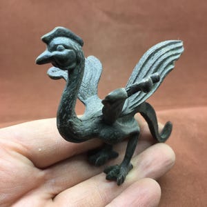 Op de afbeelding: Een kleine bronzen figuur in antieke stijl van een mythische vogel met een lange nek, gedetailleerde vleugels en een kuif. De vogel staat met zijn vleugels iets gespreid en zijn snavel open. Het beeldje wordt in een hand gehouden.