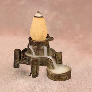 May include: A brown incense burner with a tiered design, featuring a small basin. A tan cone-shaped incense sits atop the burner, with a white and brown tip. The smoke from the incense creates a waterfall effect, adding to the aesthetic.