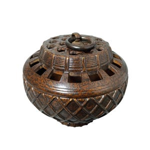 May include: A bronze-coloured container with a domed lid and a ring handle. The container has geometric cutouts and a diamond pattern on the lower section. The lid features a star-like design. The overall shape is round and bulbous, with a vintage aesthetic.