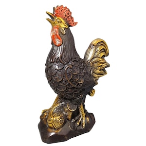 Puede incluir: Estatua de gallo de color bronce con detalles dorados. El gallo tiene una cresta y una barbilla rojas, y sus plumas están intrincadamente detalladas. Se alza sobre una base oscura, con monedas a sus pies, y su pico está abierto como si cantara.