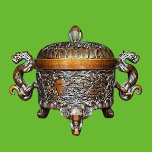 May include: A bronze-toned metal incense burner featuring an intricate relief pattern on the body, two dragon-shaped side handles, three elephant-head legs, and a removable domed lid with a perforated design and a central finial knob.