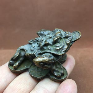 May include: A small, bronze-colored frog figurine with intricate details. The frog is depicted with a textured skin and sits atop a base. The figurine has a weathered appearance, suggesting age or a vintage style. The frog is a symbol of wealth and prosperity.