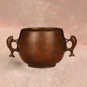 May include: A bronze-colored, small, round bowl with two ornate handles. The bowl has a wide opening and a slightly rounded base. The handles are shaped like stylized birds or animals. The background is a soft, muted pink.
