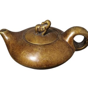 Op de afbeelding: Een bronzen theepot met een afgeronde body, een gebogen tuit en een ronde handgreep. Het deksel is bekroond met een klein olifantenbeeldje. De theepot heeft een getextureerd oppervlak en een vintage esthetiek.