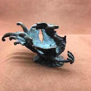 Op de afbeelding: Een bronzen sculptuur van een mythisch wezen met een gapende mond en uitgestrekte ledematen. De binnenkant van de mond heeft een contrasterende blauwgroene kleur, wat de details van het kunstwerk benadrukt.