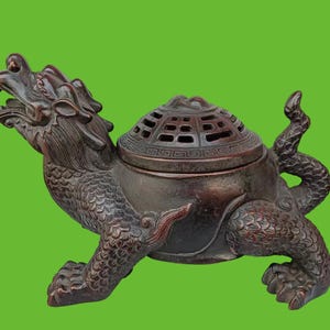 May include: A dark brown incense burner shaped like a mythical dragon with a turtle-like body. The creature features detailed scales, a wide open mouth, and a removable lid with a grid pattern. The design includes a curved tail and four legs.