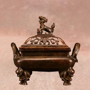 Puede incluir: Un quemador de incienso de bronce con tapa y asas decorativas. La tapa presenta un diseño calado y una pequeña estatua de una criatura mítica. El quemador descansa sobre tres patas, cada una adornada con una figura animal detallada. Una pieza clásica para la decoración del hogar.
