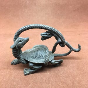 Könnte beinhalten: Eine Bronzeskulptur einer Schildkröte und einer Schlange. Die Schildkröte hat ein detailliertes Schalenmuster und ist auf allen Vieren. Die Schlange windet sich um die Schildkröte, ihr Kopf in der Nähe des Kopfes der Schildkröte. Patinierte Oberfläche.
