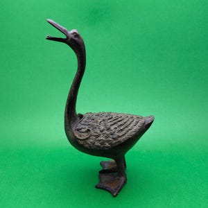 May include: A bronze-colored duck figurine with an open beak and a long, curved neck. The duck has detailed feather textures on its body and webbed feet, standing on a green background. The sculpture is a decorative item.