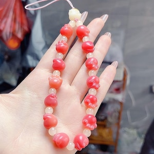 May include: A beaded necklace featuring alternating red, flower-shaped beads and smaller, round, white beads. The necklace has a pink cord and a small, white charm at the top. The beads are a vibrant red color.