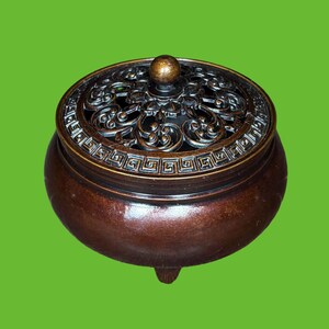 May include: A round, dark bronze-coloured metal incense burner with three small feet. The lid features an intricate, openwork filigree pattern with a central spherical knob and a decorative Greek key border around the outer edge of the lid.