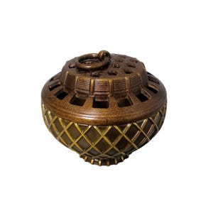 May include: A bronze-coloured decorative container with a lid and a ring handle. The lid has a series of square cutouts. The lower portion of the container features a diamond pattern with gold accents. The container is designed to hold small items.