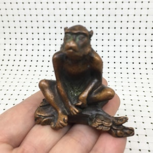 Puede incluir: Pequeña figura de mono de color bronce en posición sentada. El mono se apoya la mano en la cara, con las piernas cruzadas. La figura tiene una textura detallada y un aspecto ligeramente envejecido.