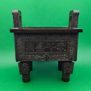 Peut inclure: Un brûleur d'encens rectangulaire en bronze foncé, orné de gravures et de caractères chinois. Il possède deux poignées et repose sur quatre pieds décoratifs. Le fond est vert uni.