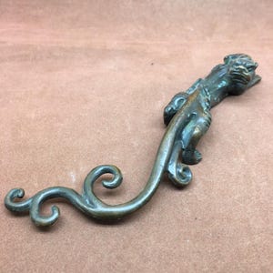 May include: A bronze-colored, stylized sculpture of a mythical creature with a long, curved tail. The creature has a detailed head and body, with a flowing tail that ends in decorative swirls. The sculpture is likely made of metal and has an aged appearance.