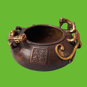 Op de afbeelding: Een donkerbruine bronzen wierookbrander met een rond lichaam, voorzien van twee goudkleurige draakvormige handvatten aan de zijkanten en een vierkant reliëfpaneel aan de voorzijde met vier Chinese karakters in een verticaal en horizontaal patroon.