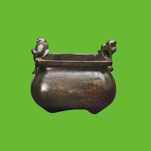 Peut inclure: Un récipient en métal de couleur bronze foncé avec une ouverture carrée et un corps arrondi. Le contenant présente deux petites figurines animales sculptées perchées sur les côtés opposés du rebord, servant de poignées décoratives.