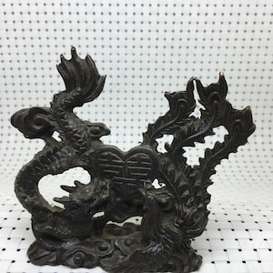 May include: A dark bronze sculpture featuring a dragon and phoenix flanking a heart-shaped emblem with Chinese characters. The detailed artwork showcases intricate patterns and textures, with the mythical creatures posed dynamically.