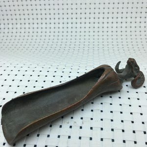 May include: A bronze-colored, elongated scoop with a ram's head detail at one end. The scoop has a curved, trough-like shape. The ram's head features curved horns and detailed facial features. The object is likely a decorative or functional item.