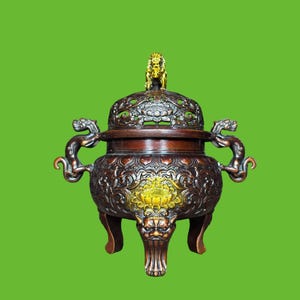 May include: A dark brown metal incense burner featuring intricate relief carvings of floral patterns and mythical creatures. The lid is topped with a gold-colored lion figure, and the body is supported by three legs with carved faces.