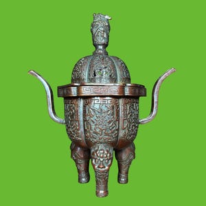 May include: A dark bronze-toned metal incense burner featuring a tripod base with three legs, each adorned with a sculpted lion-like face. The body and lid display intricate relief carvings of mythical creatures and geometric patterns, topped with a finial.