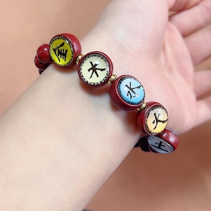 May include: A red beaded bracelet with circular charms featuring Chinese characters in yellow, black, and white. The bracelet is worn on a wrist and has a mix of red and gold accents.
