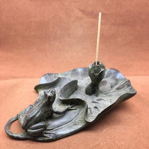 Puede incluir: Un porta incienso de bronce con una rana sobre una hoja de nenúfar. El soporte tiene un diseño detallado con una rana a la izquierda y una flor de loto con una varilla de incienso en el centro. La hoja de nenúfar está detallada con venas.
