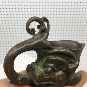 May include: A bronze-colored, small cup with a handle. The cup is adorned with a detailed, mythical creature design on the front, featuring a face with prominent features. The handle has a unique, curved shape.