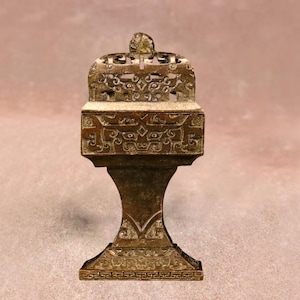 May include: A bronze-coloured incense burner with intricate carvings and a decorative lid. The burner features a rectangular body with detailed patterns and sits on a pedestal base. The design suggests an Asian cultural influence.