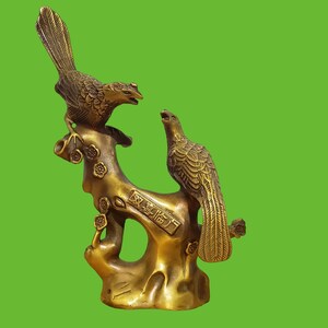 May include: A brass sculpture featuring two birds perched on a textured tree branch with small flower details. The branch includes an engraved rectangular plaque with Chinese characters. The metallic surface has a polished, golden-brown finish.