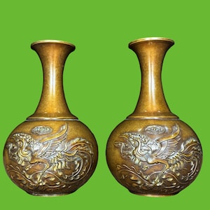 Pode incluir: Um par de vasos a condizer em tom de bronze com corpo bulboso e pescoço fino e alargado. Cada vaso apresenta um relevo detalhado de uma ave mítica com as asas abertas, rodeado por padrões estilizados de nuvens e folhagem em metal.