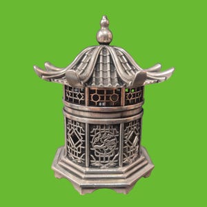 May include: A silver-toned metal lantern featuring a multi-tiered roof with curved eaves and a finial top. The cylindrical body displays intricate lattice panels, including a circular cutout with a relief design of a tree, set on a base.