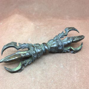 May include: A bronze vajra, a ritual object with three curved prongs at each end. The central handle has detailed embossed patterns. The metal has an aged look, with traces of green patina in the grooves.