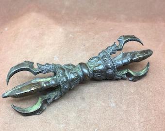 May include: A bronze-colored vajra, a ritual object with three curved prongs on each end. The central handle features intricate embossed details. The metal has a slightly aged appearance, with hints of green patina in the crevices.