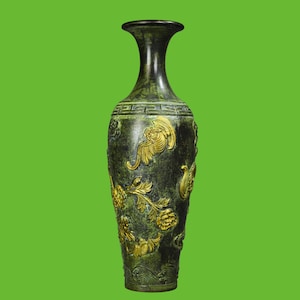 May include: A tall, slender vase with a dark, weathered green patina finish. The surface is decorated with raised, gold-colored relief carvings of floral motifs, swirling clouds, and a geometric Greek key pattern around the neck.