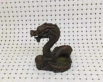 May include: A bronze-colored dragon figurine with an open mouth and a curved body. The dragon is detailed with scales and a textured surface. The figurine is a decorative item, possibly for a desk or shelf.