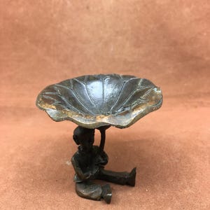 Puede incluir: Una escultura de bronce de una figura sentada bajo una gran hoja de loto. La figura está detallada, con ropa y rasgos faciales visibles. La hoja tiene vetas texturizadas y una apariencia desgastada, lo que sugiere un acabado antiguo.