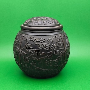 Könnte beinhalten: Eine dunkelbraune, runde Dose mit Deckel, mit aufwendigen Schnitzereien chinesischer Schriftzeichen, Bambus und Wolken. Die Dose hat eine glatte, matte Oberfläche und steht vor einem einfarbigen grünen Hintergrund. Der Deckel hat ebenfalls geschnitzte Details.